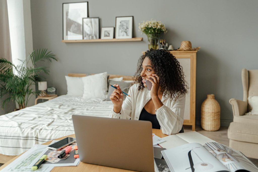 A woman in a home office multitasking with a laptop and phone call, surrounded by documents.