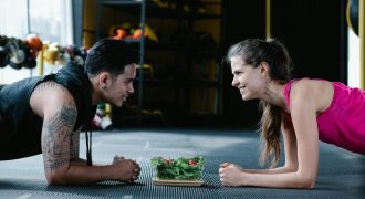 Two people planking face-to-face in a gym with a salad between them, smiling and engaging in a fitness activity.