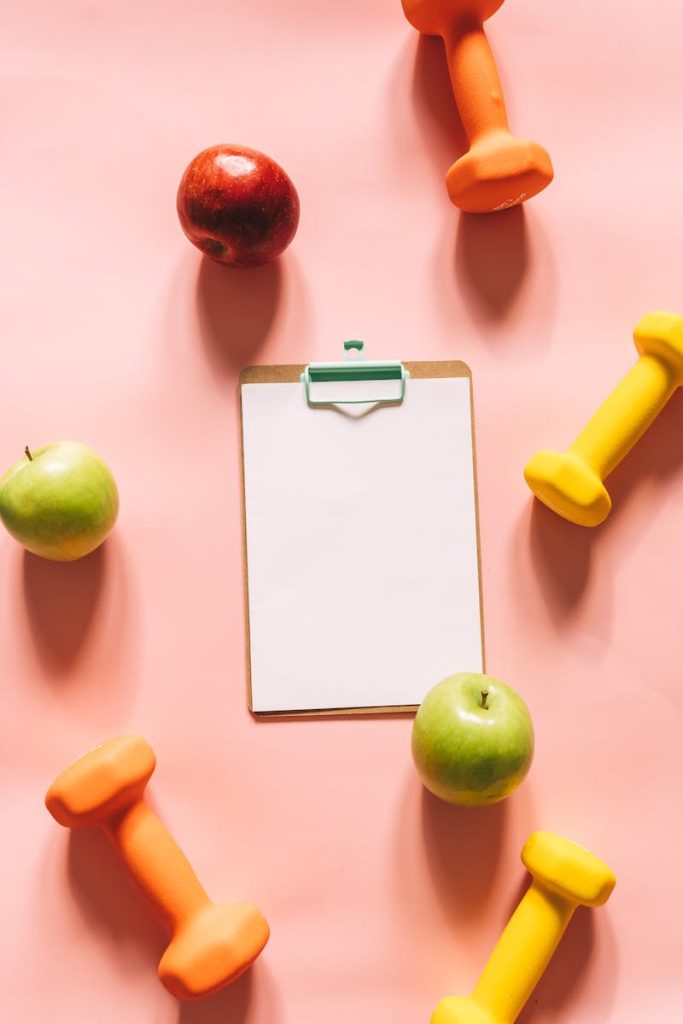 Top view of colorful apples, dumbbells, and clipboard on a pink background, ideal for health and fitness themes.
